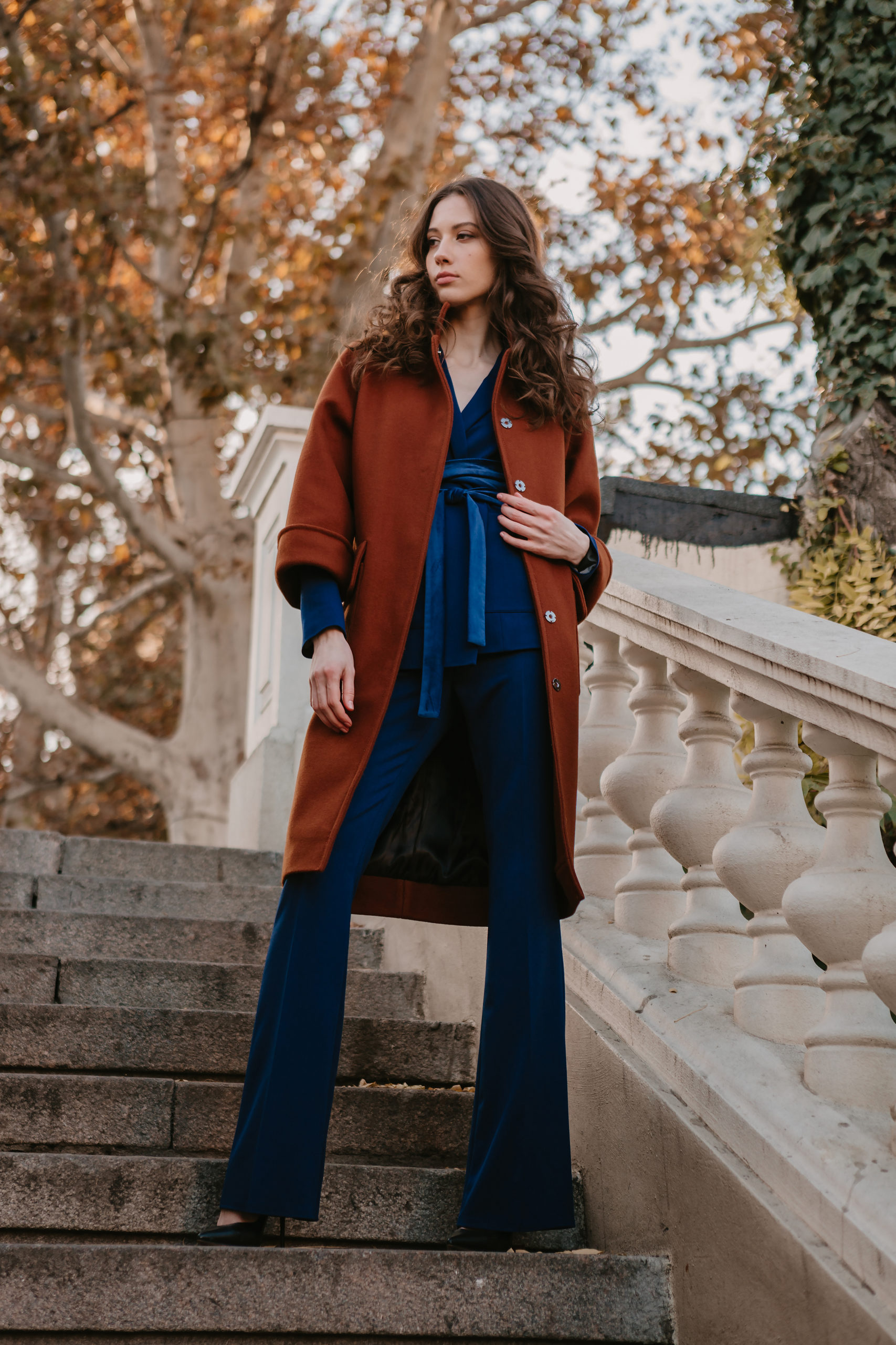 beautiful stylish smiling skinny woman with curly hair walking in street stairs dressed in warm brown coat and blue suit, autumn trendy fashion street style